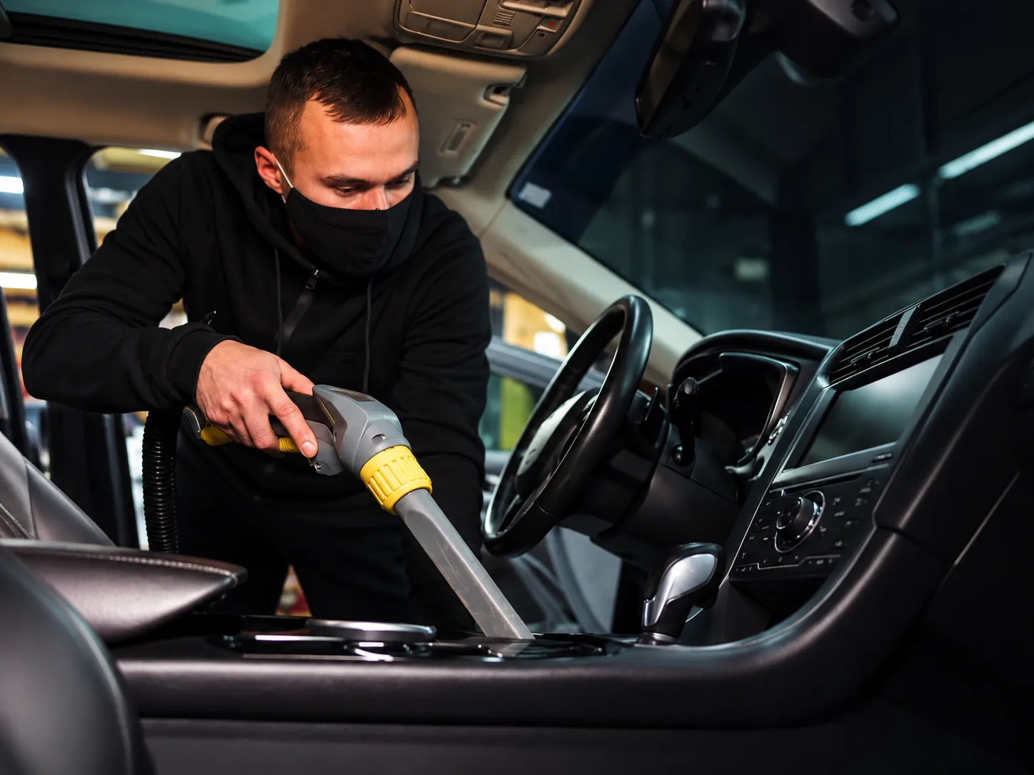 Técnico aspirando o interior do carro durante a higienização, luvas azuis, iluminação de oficina, foco no detalhe do estofado